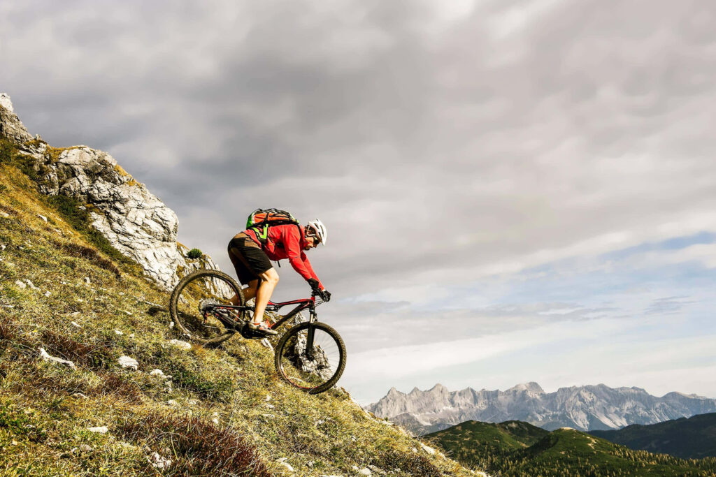 Cyclist in red jacket rides rugged mountain trail under cloudy sky with distant peaks background Wallpaper