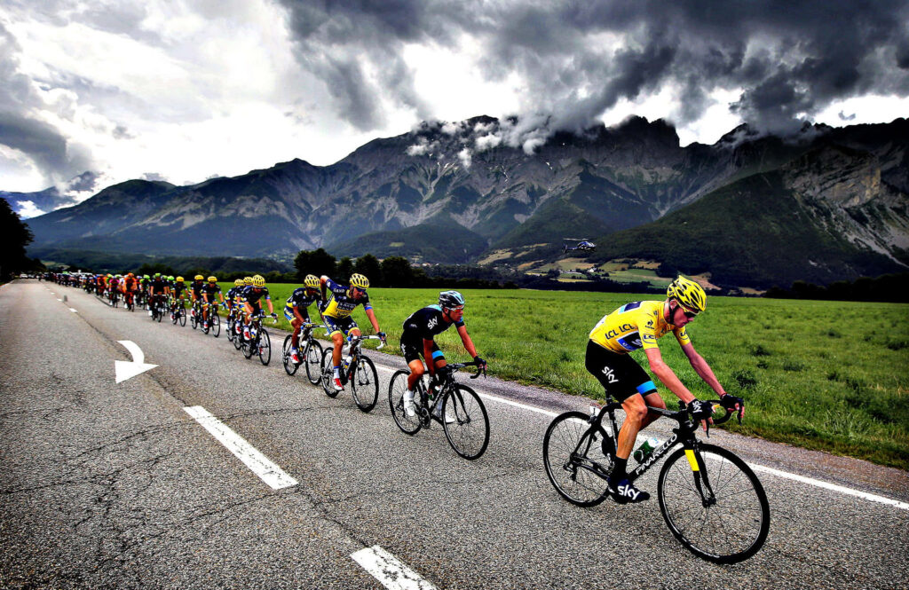 Cyclists racing on winding road with green fields and mountains under dramatic cloudy sky wallpaper