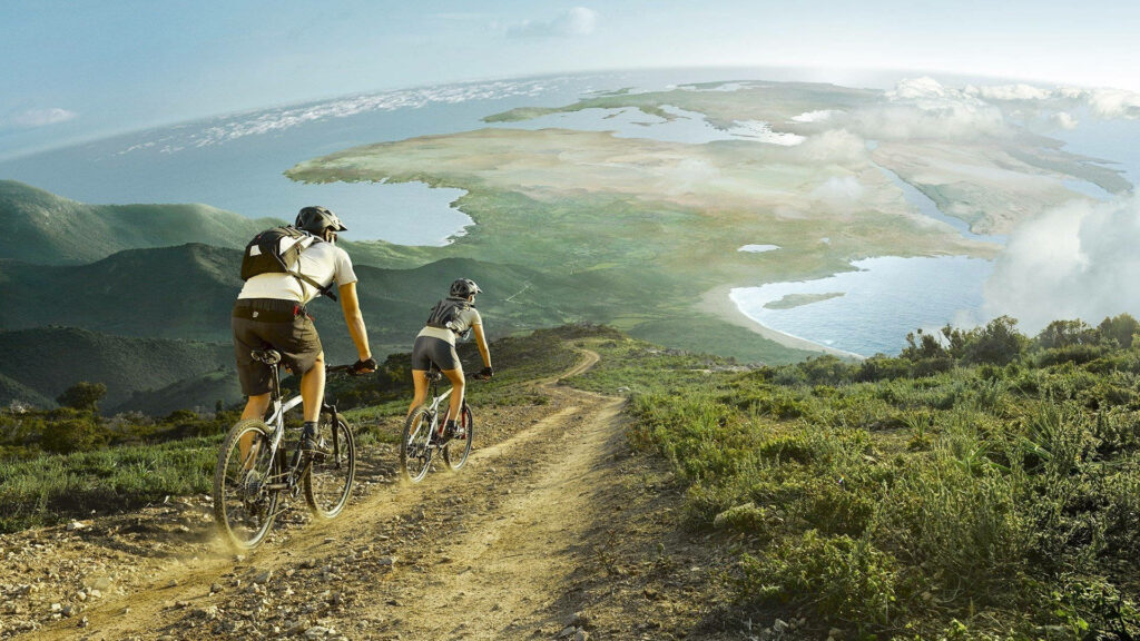Two cyclists ride winding hillside trail under bright sky with panoramic landscape views wallpaper
