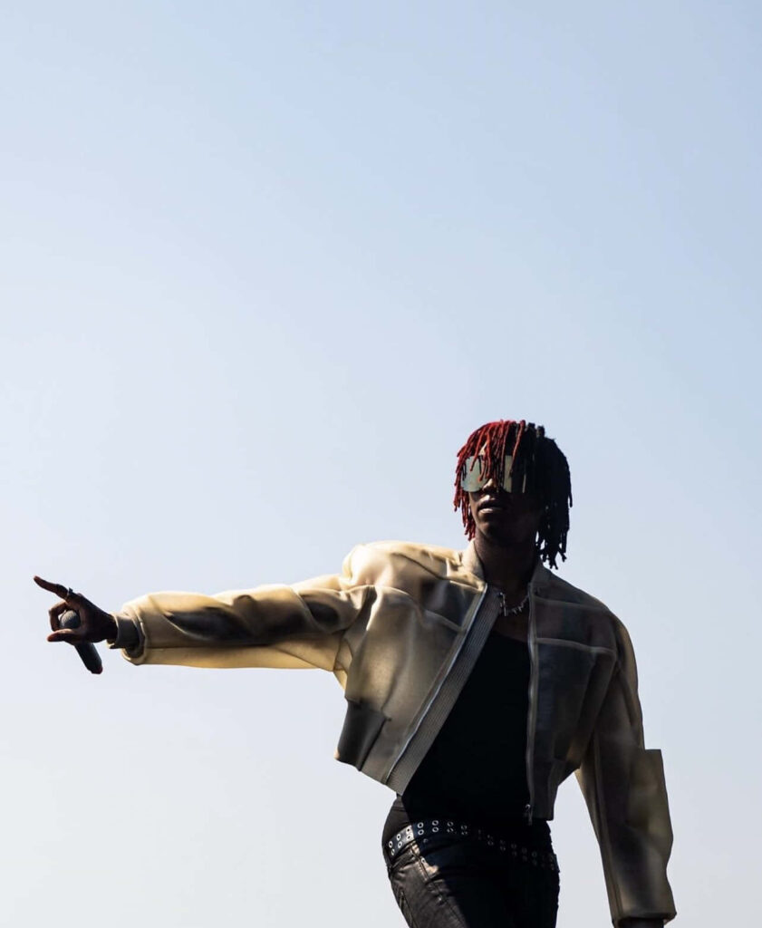 DC The Don with red dreadlocks bold fashion jacket and studded belt against clear sky background Wallpaper