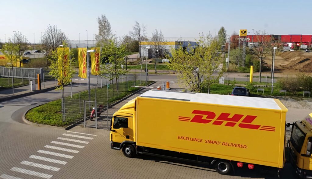 Bright yellow DHL truck with red branding driving through modern logistics facility outdoors Wallpaper