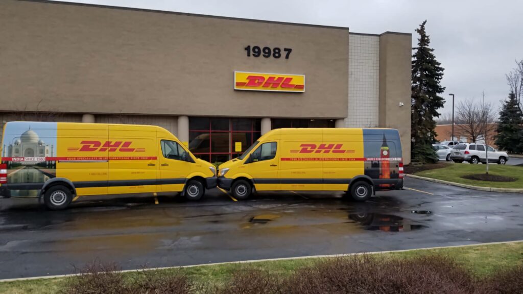 Two yellow DHL vans parked outside beige DHL building with wet pavement and bold red logos Wallpaper