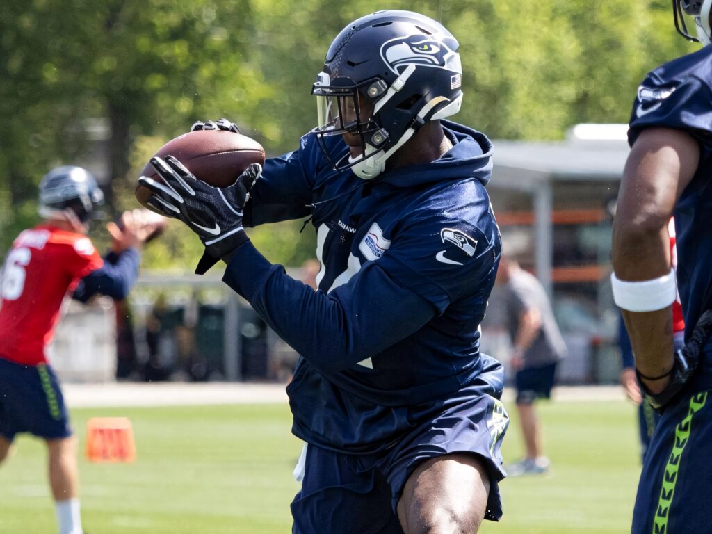 DK Metcalf in Seahawks training catching football wearing navy jacket and helmet on green field Wallpaper