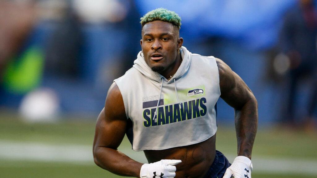 DK Metcalf mid-workout in Seahawks hoodie showcasing athletic physique on vibrant football field background Wallpaper