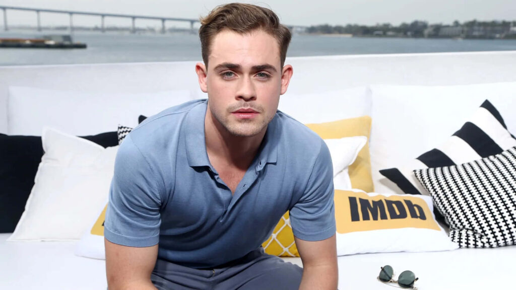 Dacre Montgomery in light blue polo seated on modern outdoor sofa with waterfront and bridge view Wallpaper