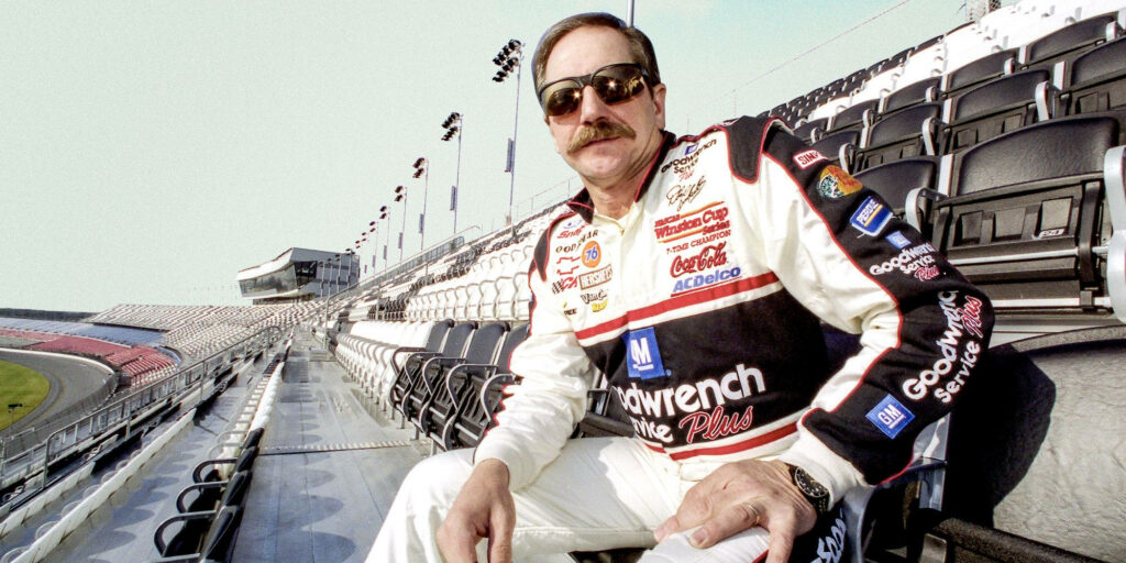 Dale Earnhardt NASCAR driver portrait in racing suit with sunglasses at sunlit racetrack grandstand Wallpaper