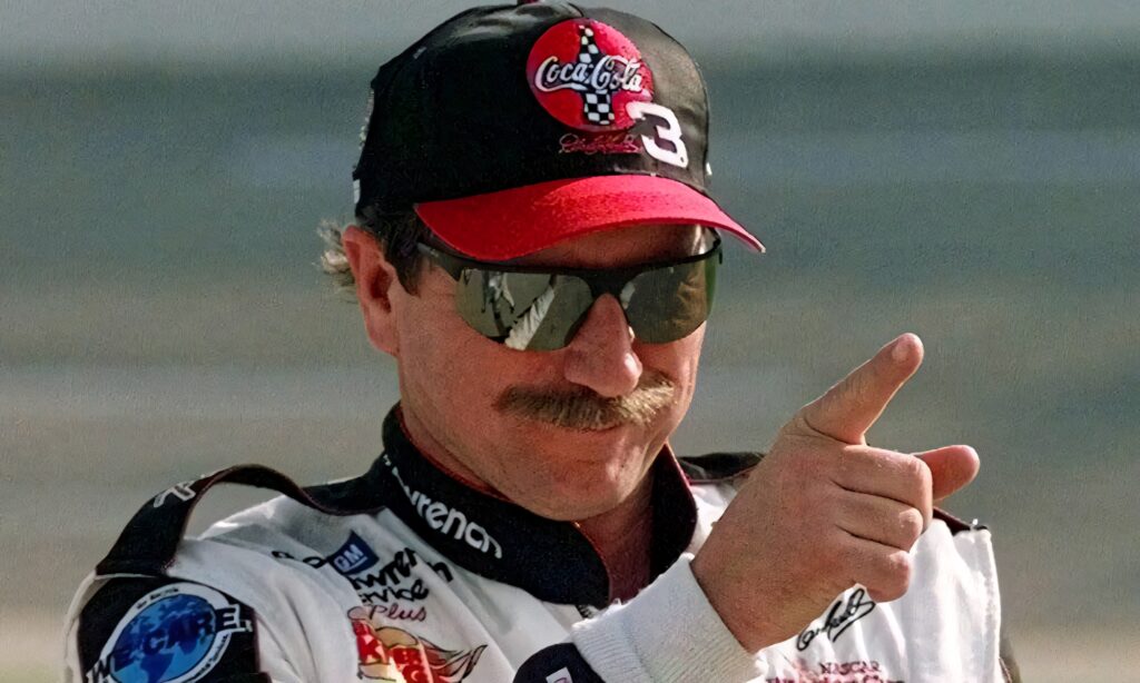 Dale Earnhardt in iconic racing suit and sunglasses pointing confidently with soft daylight background Wallpaper