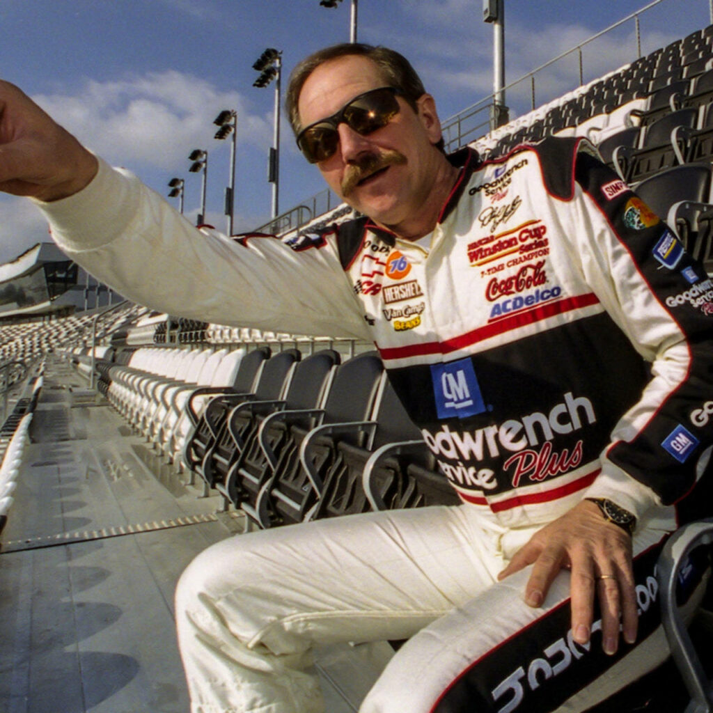 Dale Earnhardt in NASCAR racing suit sitting on stadium bench with empty seats in sunlight Wallpaper