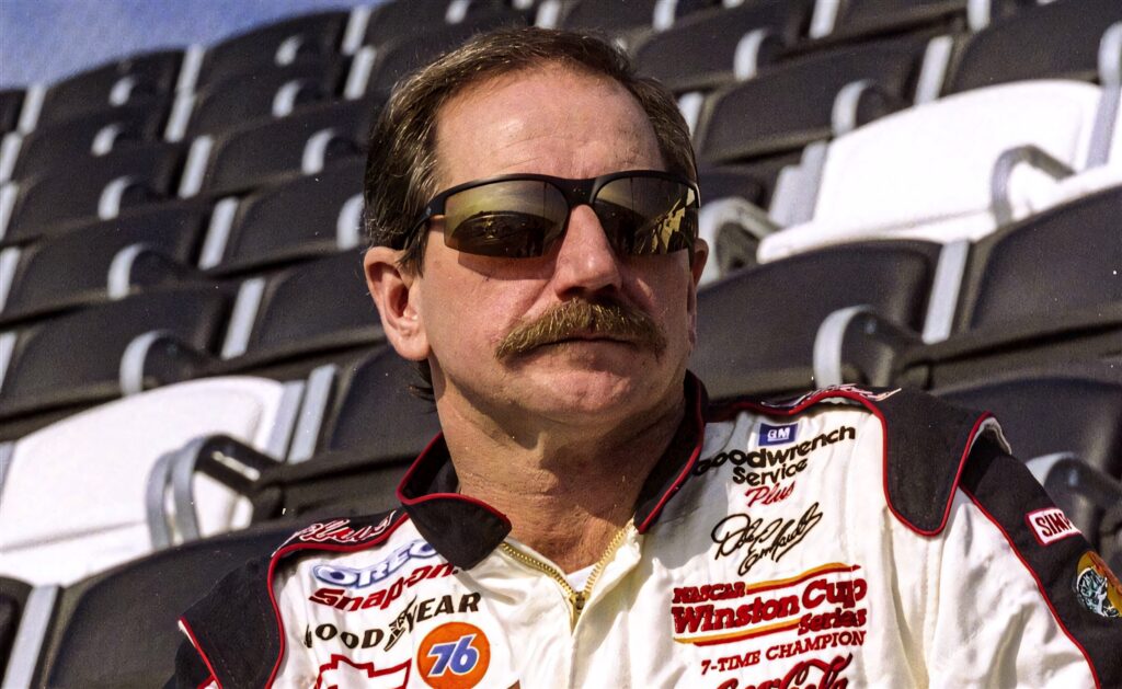 Dale Earnhardt in racing suit and sunglasses sitting confidently in empty stadium stands wallpaper