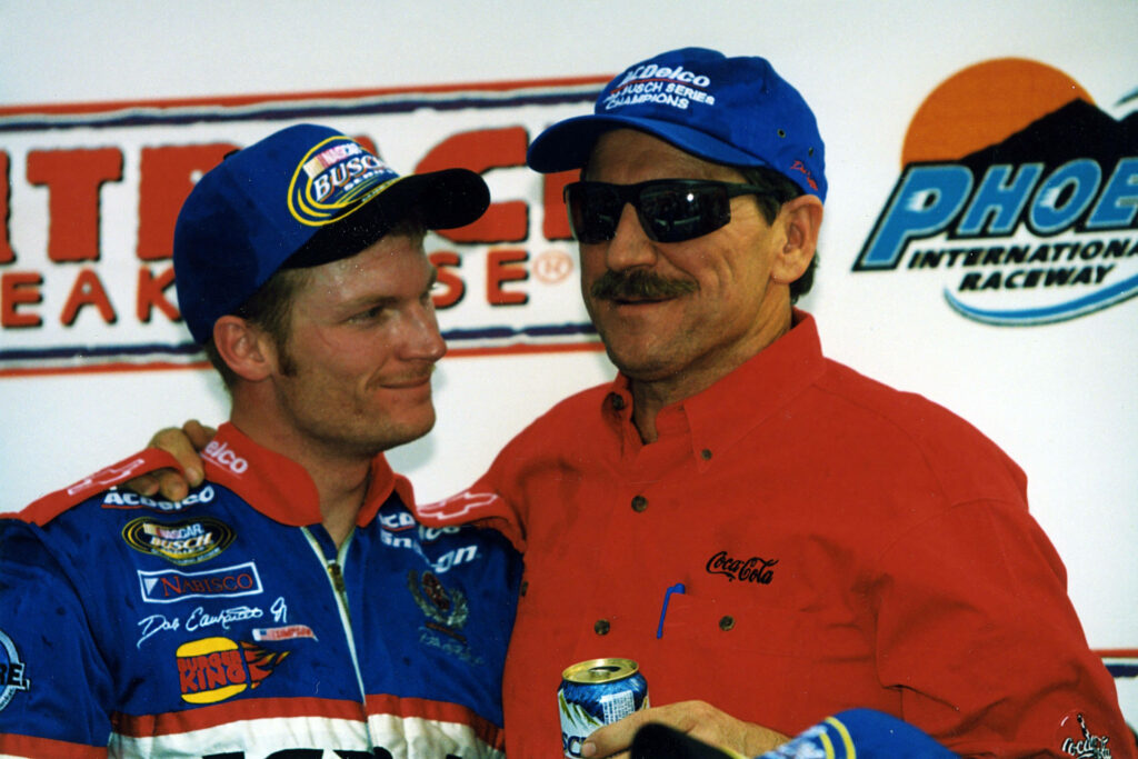 NASCAR legends Dale Earnhardt Sr and Jr in racing suits share a proud moment vibrant backdrop Wallpaper