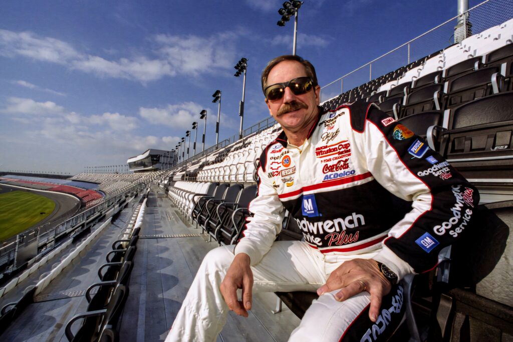 Dale Earnhardt in iconic Goodwrench racing suit stands in empty NASCAR stadium under bright sky Wallpaper