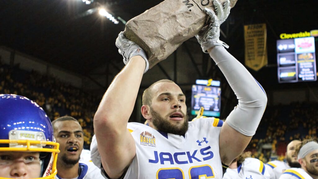 Dallas Goedert in white blue Jacks uniform celebrates with paper bag trophy in vibrant stadium Wallpaper