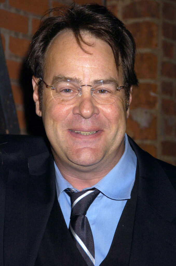 Dan Aykroyd in dark suit and striped tie smiling against rustic red brick wall wallpaper background