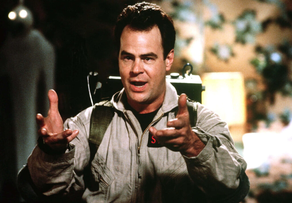 Dan Aykroyd as Ray Stantz in beige Ghostbusters jumpsuit with proton pack on wallpaper background