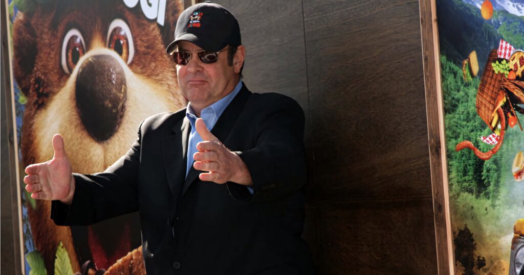 Dan Aykroyd in dark suit and sunglasses posing before vibrant animated wallpaper background