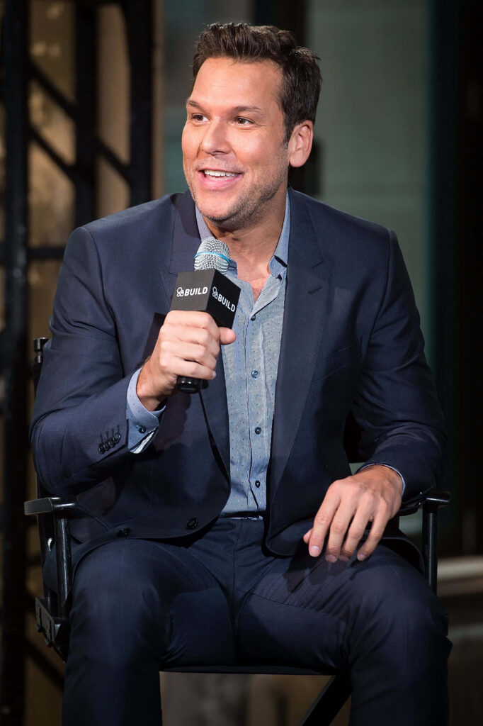 Dane Cook smiling in navy suit holding microphone during interview with blurred background wallpaper