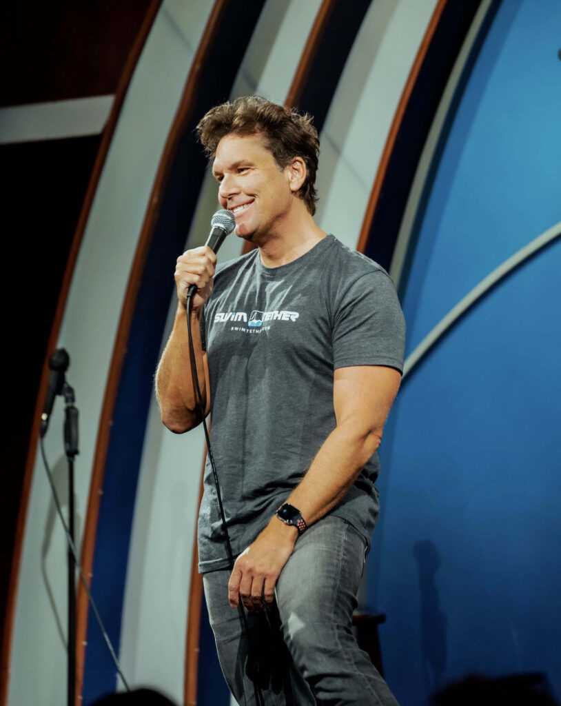 Dane Cook performing live stand-up comedy on stage with blue and white arc background wallpaper