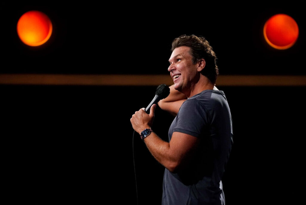 Dane Cook performing stand-up comedy on stage with microphone under warm spotlights wallpaper
