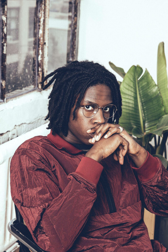 Daniel Caesar portrait in maroon jacket and glasses against rustic window with soft lighting wallpaper
