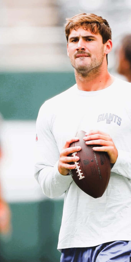 Daniel Jones focused with football in hand wearing Giants sweatshirt during outdoor practice session Wallpaper