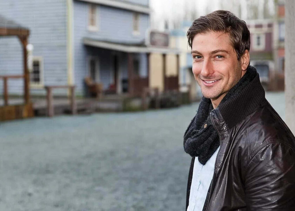 Daniel Lissing in leather jacket and scarf smiles outdoors by pastel wooden buildings in rustic town Wallpaper