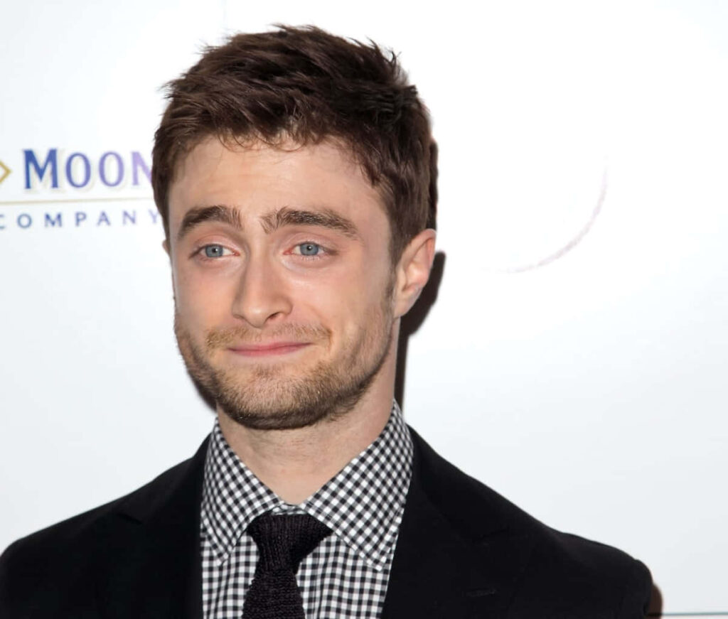 Daniel Radcliffe in black suit and checkered shirt with blue eyes on bright minimal wallpaper background
