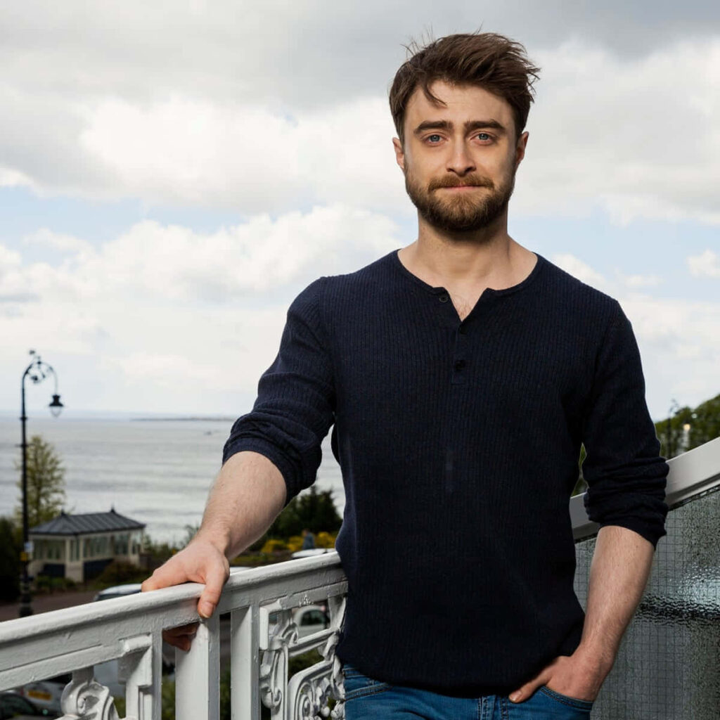 Daniel Radcliffe in dark blue sweater on balcony with ocean view soft clouds natural light wallpaper