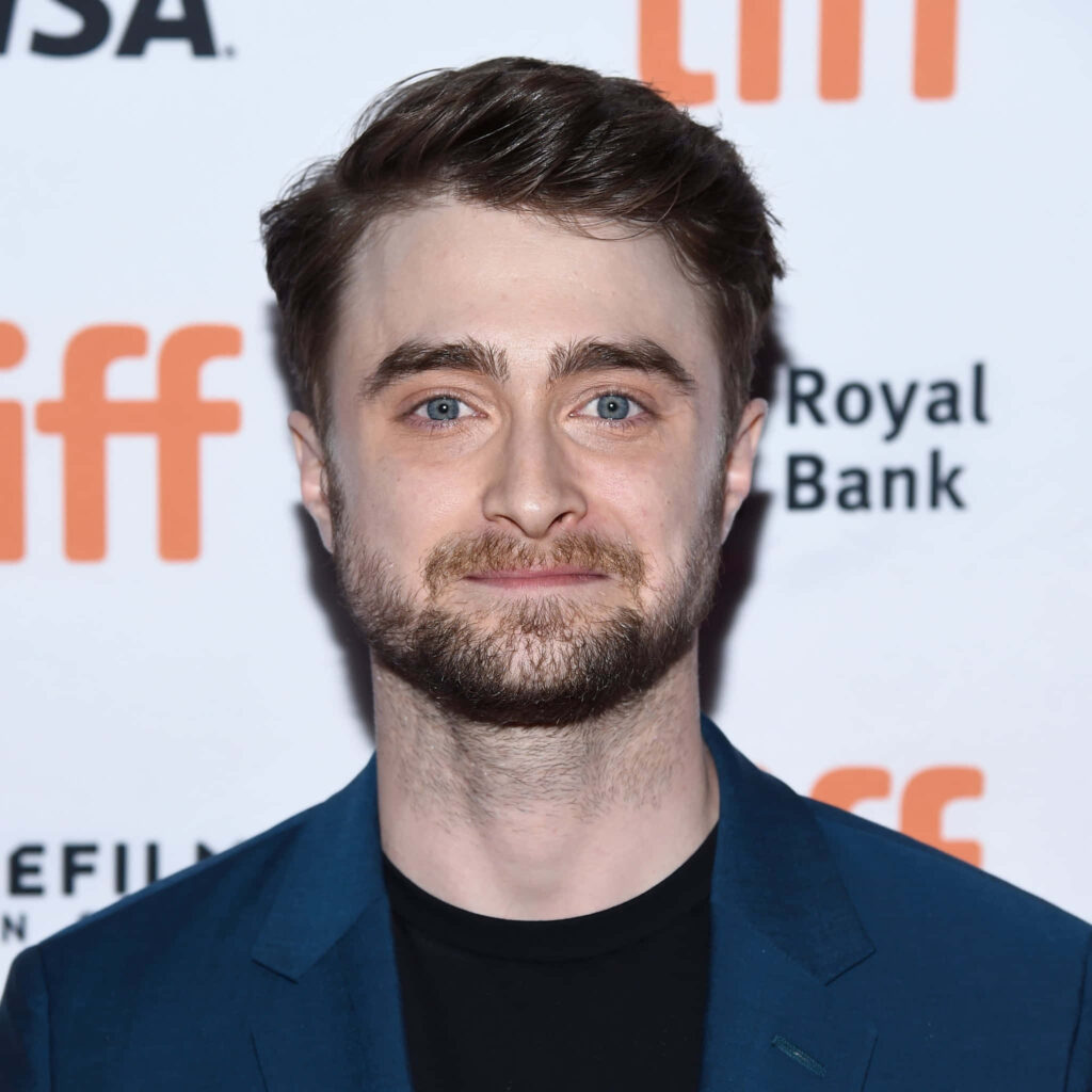 Daniel Radcliffe in dark blue suit smiling against white backdrop with event branding wallpaper