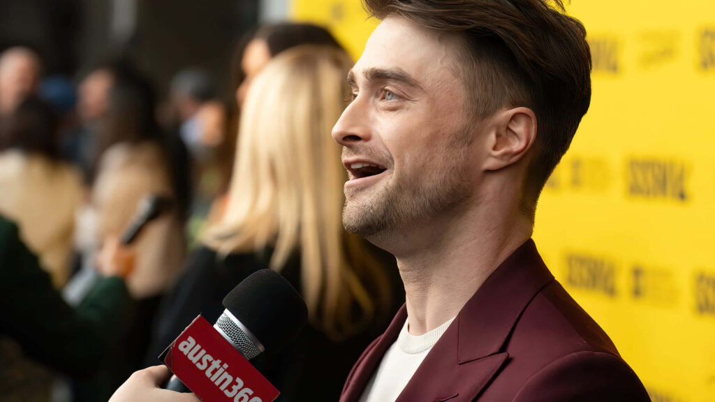 Daniel Radcliffe smiling in maroon suit during interview at event with bright yellow backdrop Wallpaper