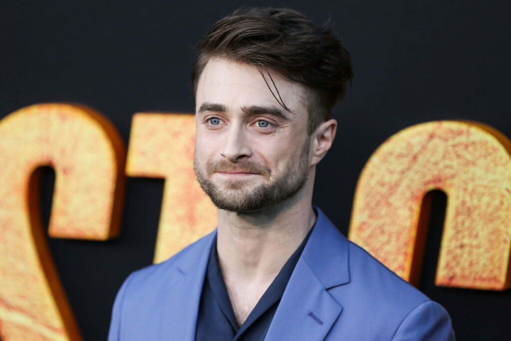 Daniel Radcliffe in blue suit with confident gaze against bold golden letter wallpaper background