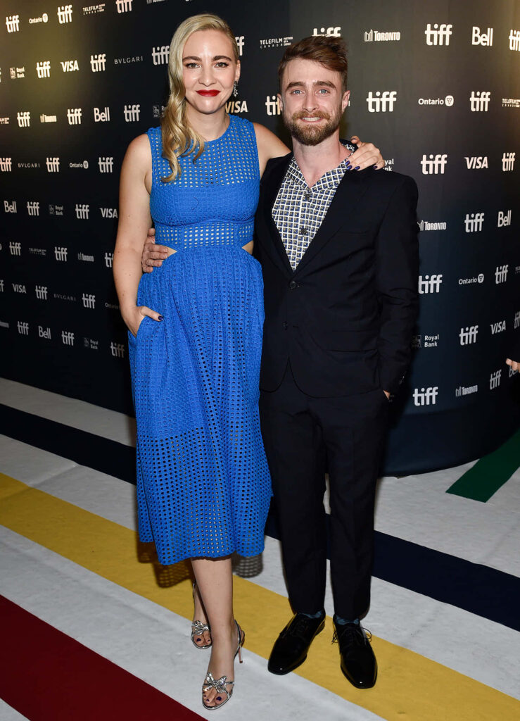 Daniel Radcliffe and Erin Darke pose on red carpet blue dress dark suit event sponsor logos background Wallpaper