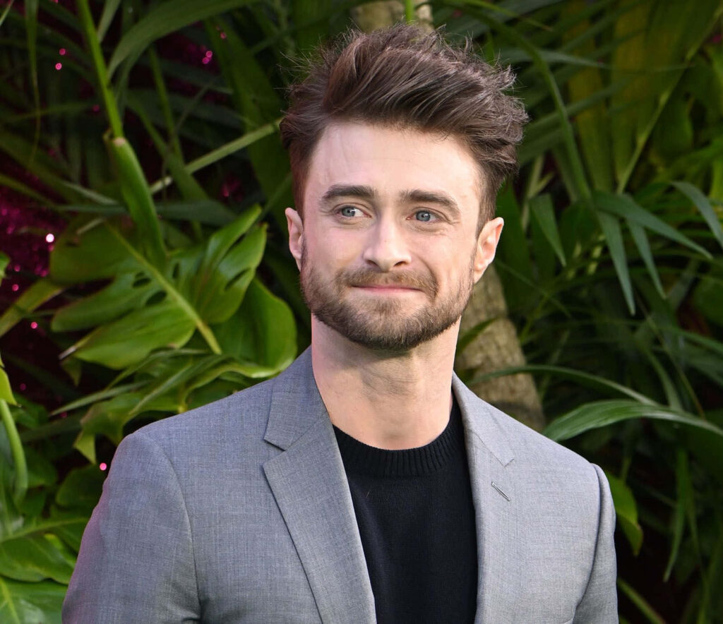 Daniel Radcliffe in sharp gray suit and black shirt standing confidently against lush green foliage Wallpaper
