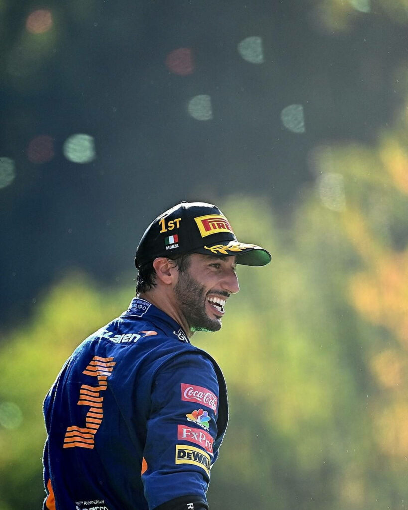 Daniel Ricciardo in racing suit and cap on sunlit Formula 1 track with green yellow bokeh background Wallpaper