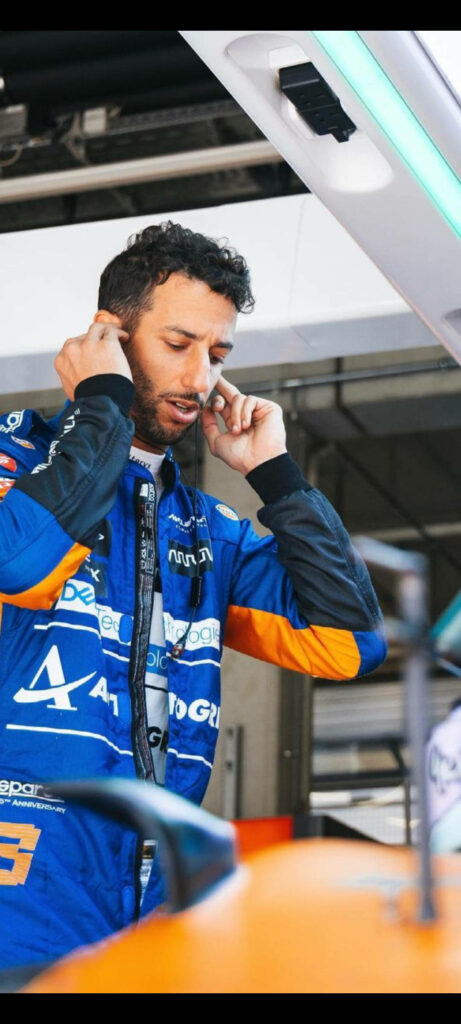 Daniel Ricciardo in blue orange racing suit puts in earplugs in motorsport garage focused preparation Wallpaper