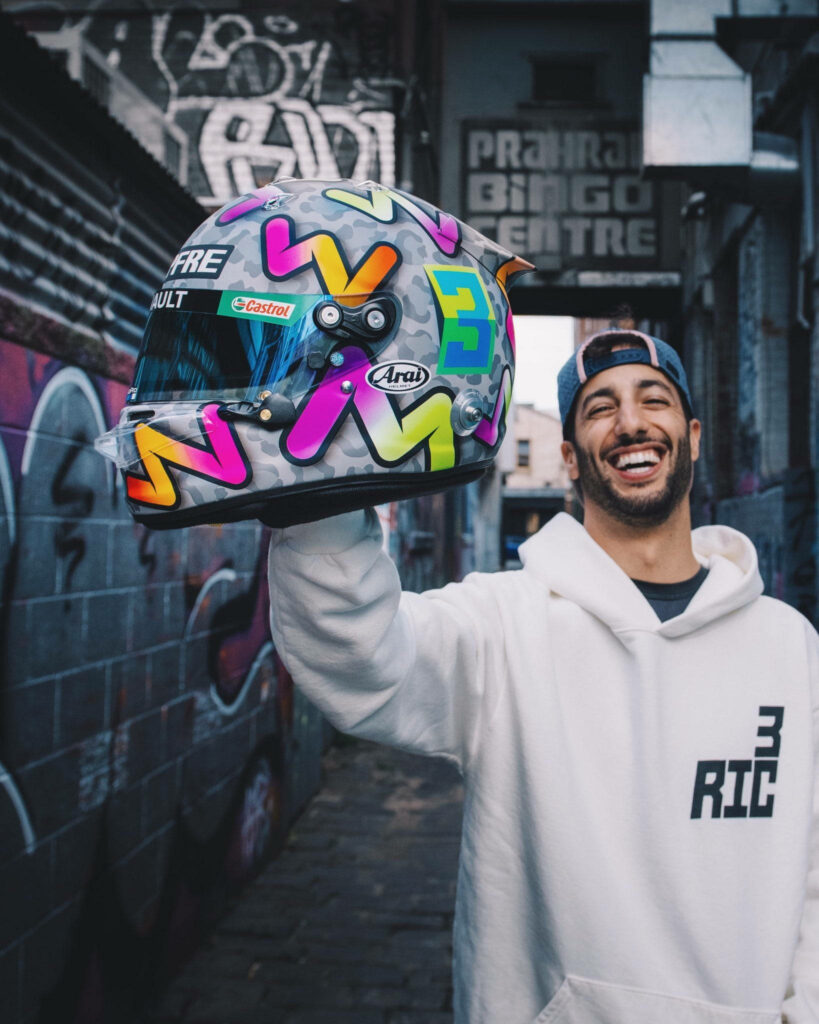 Daniel Ricciardo in white hoodie holding colorful racing helmet in vibrant graffiti alleyway wallpaper