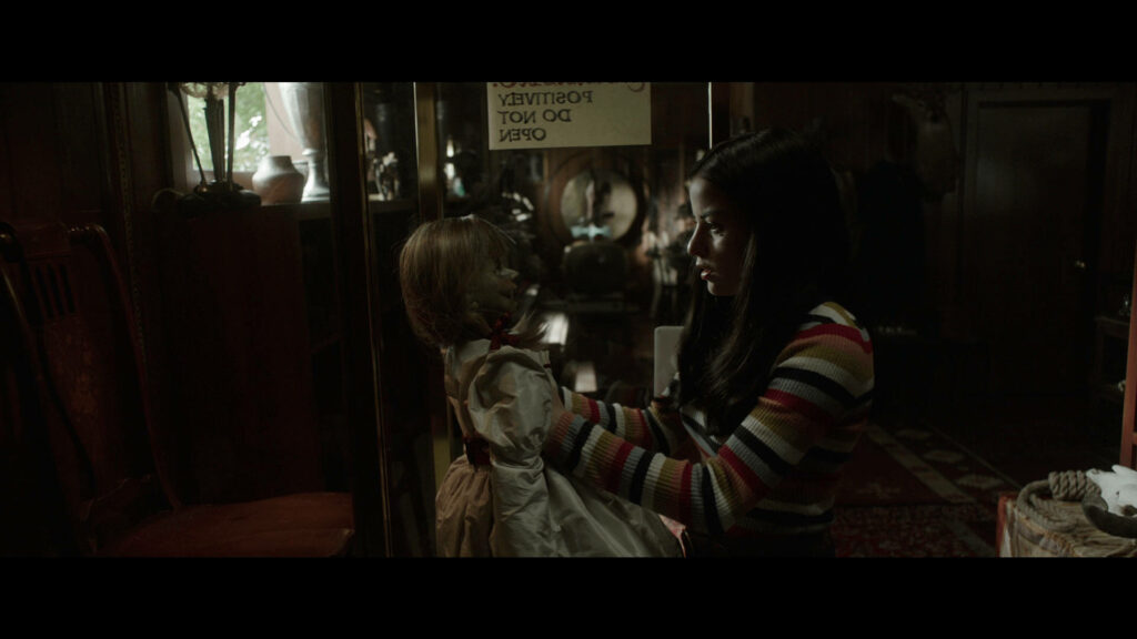 Daniela Rios holds eerie Annabelle doll in glass case in dim antique room with wallpaper background