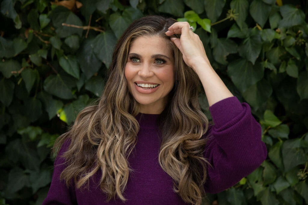 Danielle Fishel smiling in purple sweater with wavy hair against vibrant green leafy background Wallpaper