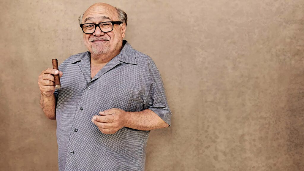 Danny DeVito in patterned shirt and glasses holding cigar against textured beige wallpaper background