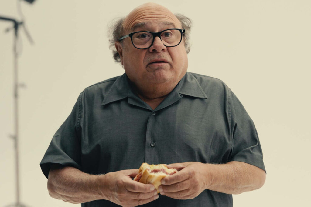 Danny DeVito in dark shirt holding sandwich with playful expression on light background wallpaper