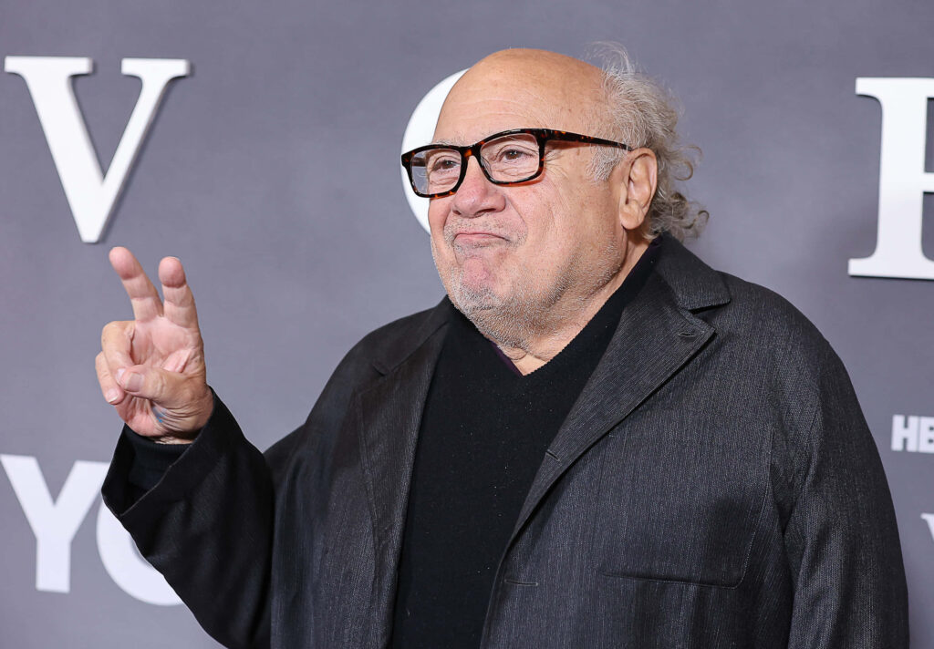 Danny DeVito in dark jacket and glasses giving playful peace sign against grey backdrop wallpaper