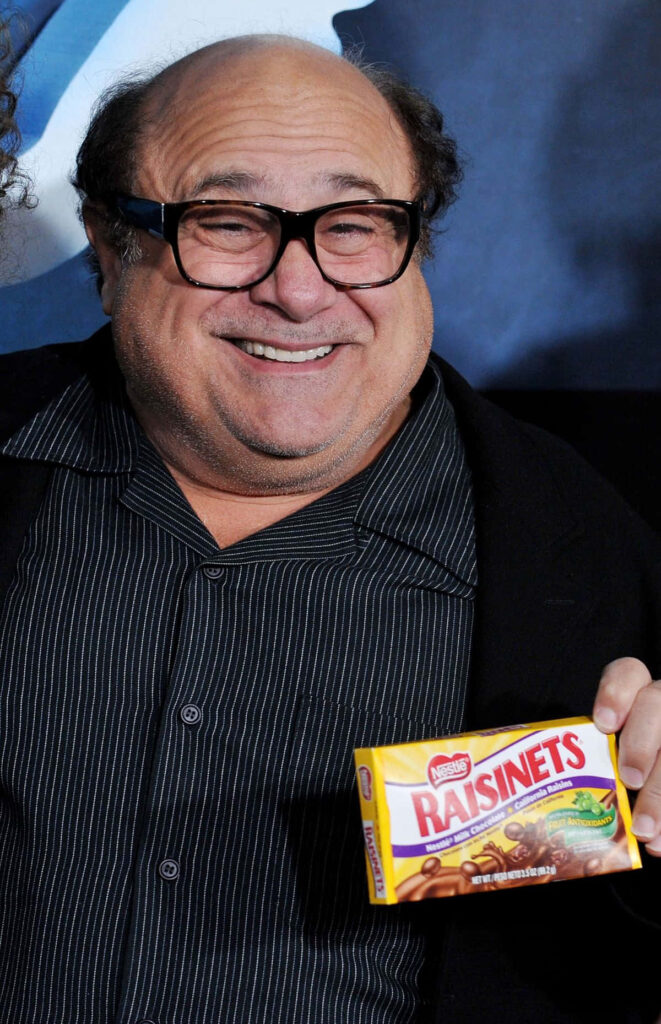 Danny DeVito smiling holding Raisinets box wearing striped shirt and signature glasses on dark background Wallpaper