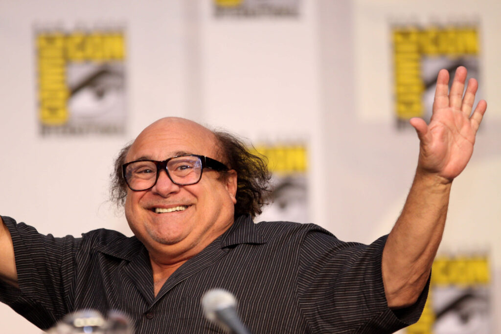 Danny DeVito smiling and raising arms at Comic-Con event in casual striped shirt with warm lighting Wallpaper
