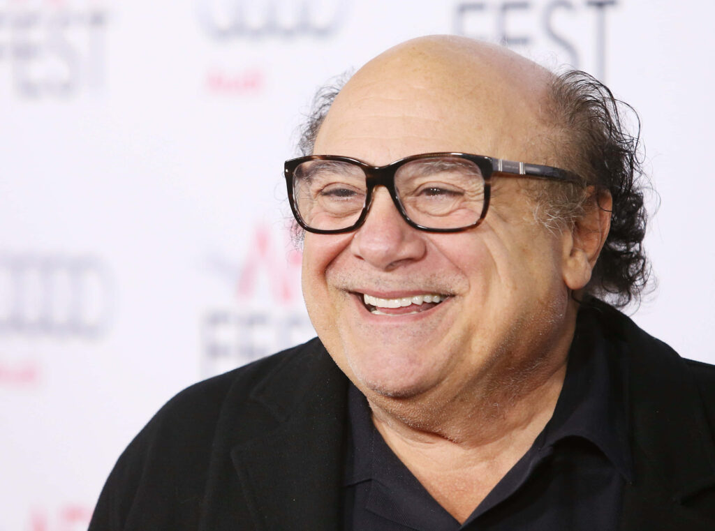 Danny DeVito smiling warmly in vibrant wallpaper with event logos and stylish contemporary look