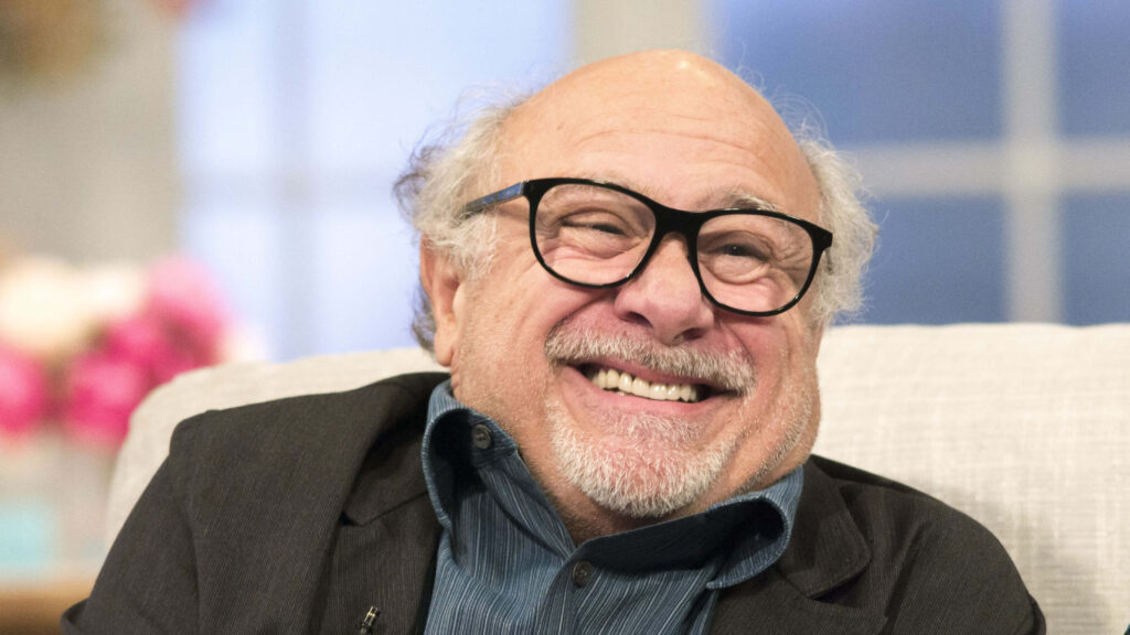 Danny DeVito smiling warmly on light couch wearing dark jacket and blue shirt pastel background wallpaper