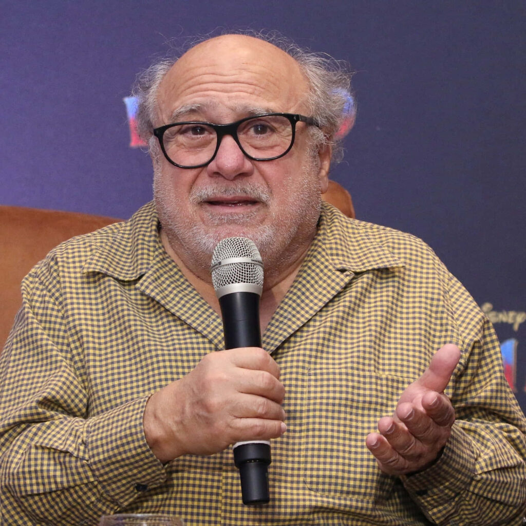 Danny DeVito speaking with microphone wearing checkered shirt on dark gradient wallpaper background