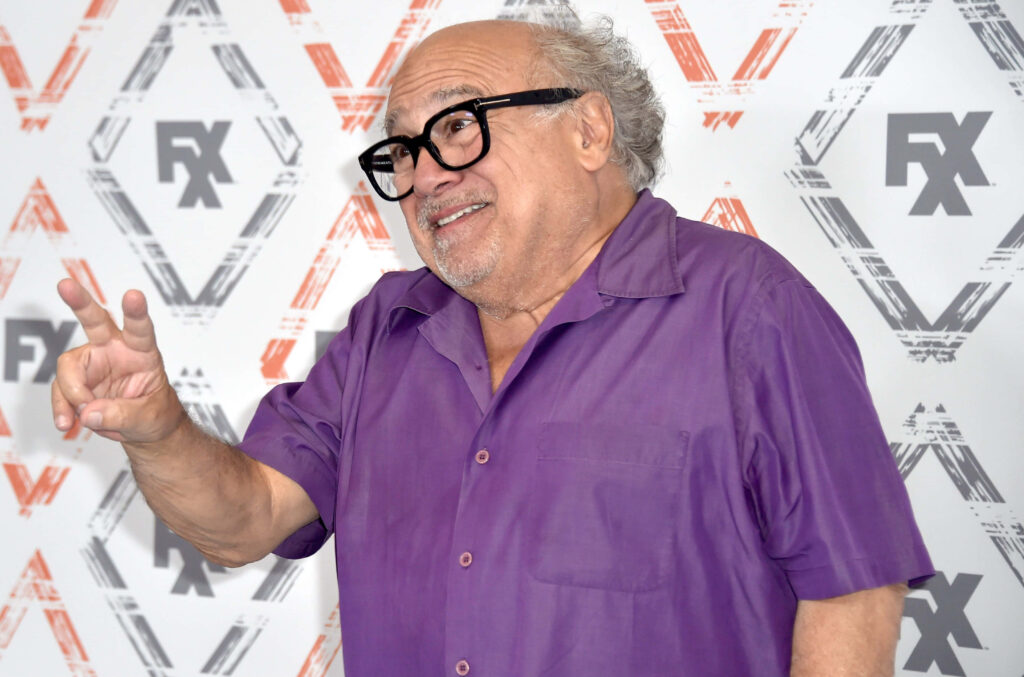 Danny Devito in vibrant purple shirt and black glasses smiling against patterned FX backdrop wallpaper