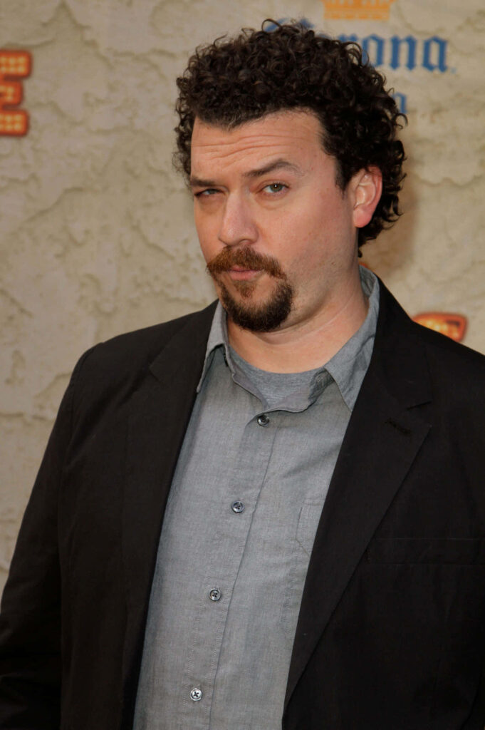 Danny McBride in casual gray shirt black blazer with curly hair against beige wall wallpaper background