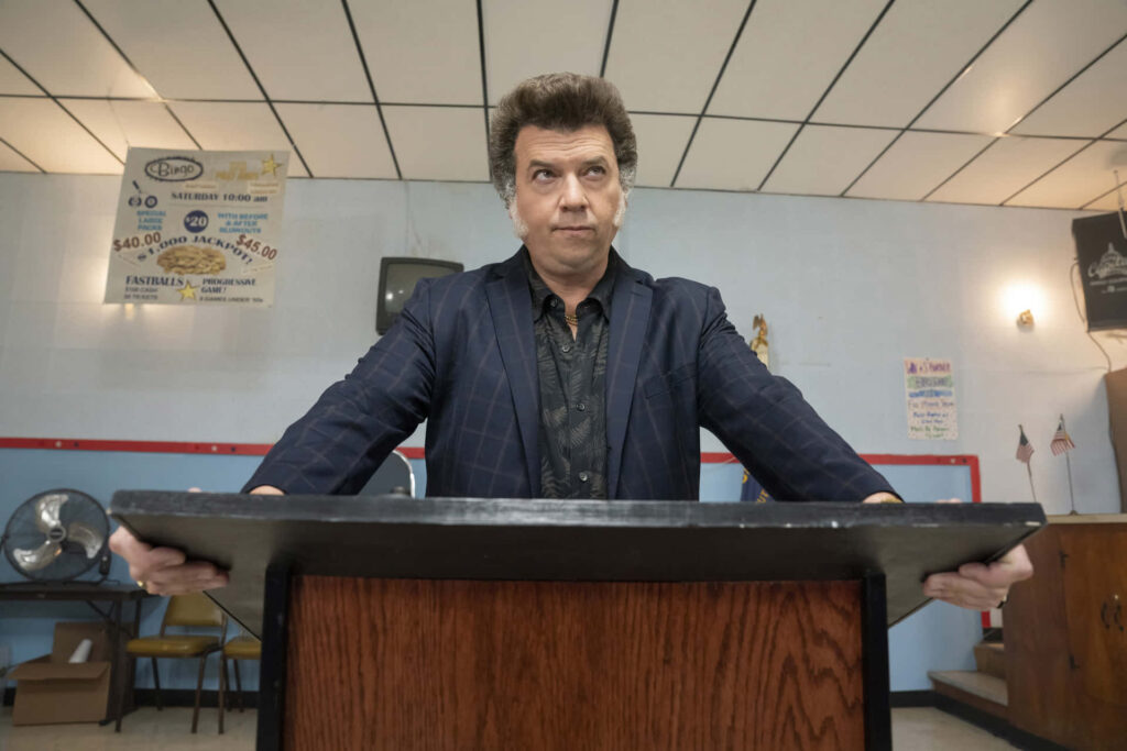 Danny McBride at wooden podium in bright room with blue red wallpaper comedic TV film setting