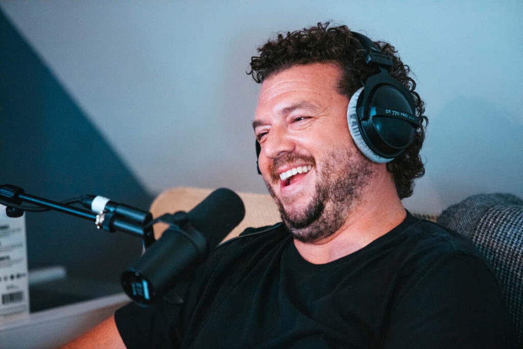Danny McBride laughing with headphones in studio podcast setting blue lighting vibrant wallpaper