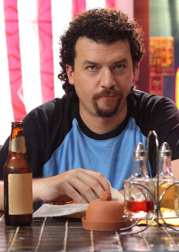 Danny McBride with curly hair and goatee at table enjoying meal with colorful wallpaper background
